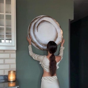 May include: A large, round decorative wall plate with a white center and a gold-colored, swirling edge. The plate is being held against a muted green wall. The plate's design resembles a stylized abstract shape. The overall aesthetic is modern and minimalist.