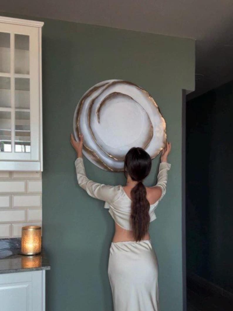 May include: A large, round decorative wall plate with a white center and a gold-colored, swirling edge. The plate is being held against a muted green wall. The plate's design resembles a stylized abstract shape. The overall aesthetic is modern and minimalist.