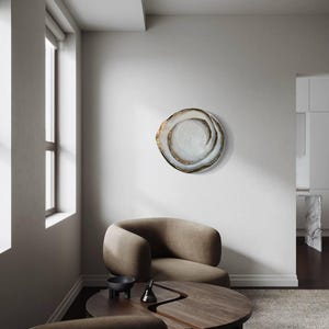 May include: A decorative wall art piece with a swirling, organic design. The artwork features a light-colored center with a dark, textured outer edge. The piece is mounted on a white wall in a modern living space, with a brown armchair and wooden table.