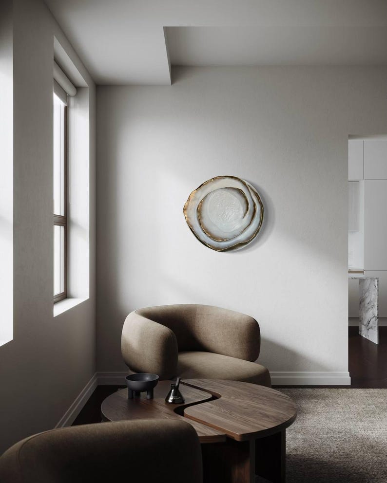 May include: A decorative wall art piece with a swirling, organic design. The artwork features a light-colored center with a dark, textured outer edge. The piece is mounted on a white wall in a modern living space, with a brown armchair and wooden table.