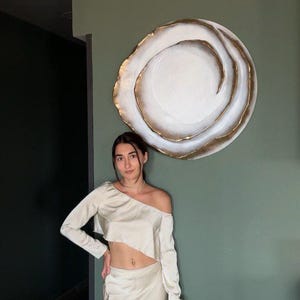 May include: A cream-colored two-piece outfit featuring a long-sleeved, off-the-shoulder crop top and a matching skirt. A large, circular wall art piece with a white center and gold trim hangs on a green wall.
