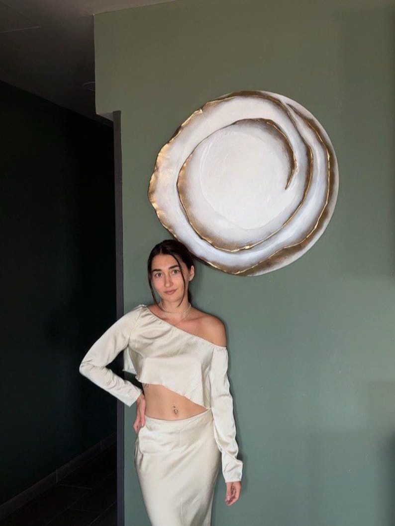 May include: A cream-colored two-piece outfit featuring a long-sleeved, off-the-shoulder crop top and a matching skirt. A large, circular wall art piece with a white center and gold trim hangs on a green wall.