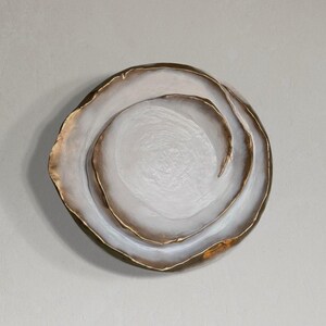 May include: A decorative, round, white and gold-rimmed plate with a textured surface. The plate has a layered design with a gold accent on the outer edge and inner swirls.