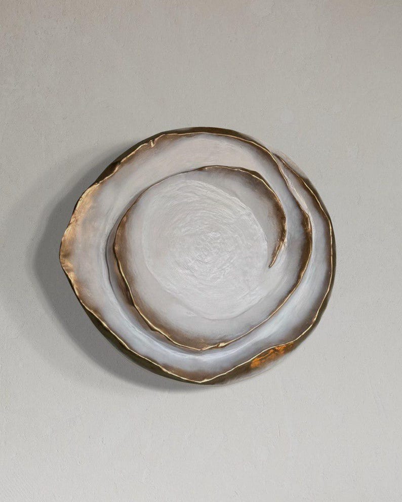 May include: A decorative, round, white and gold-rimmed plate with a textured surface. The plate has a layered design with a gold accent on the outer edge and inner swirls.