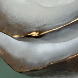 May include: Close-up of a decorative, abstract ceramic bowl. The bowl features a flowing design with a metallic gold rim and accents against a matte gray and white surface. The bowl's shape is organic and modern.