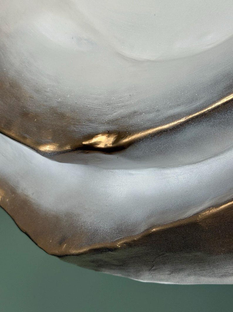 May include: Close-up of a decorative, abstract ceramic bowl. The bowl features a flowing design with a metallic gold rim and accents against a matte gray and white surface. The bowl's shape is organic and modern.