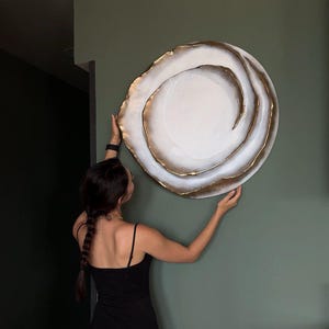 May include: A large, round wall art piece with a white center and gold-colored, layered edges. The artwork is being held against a muted green wall. The piece has a spiral design and a textured appearance.