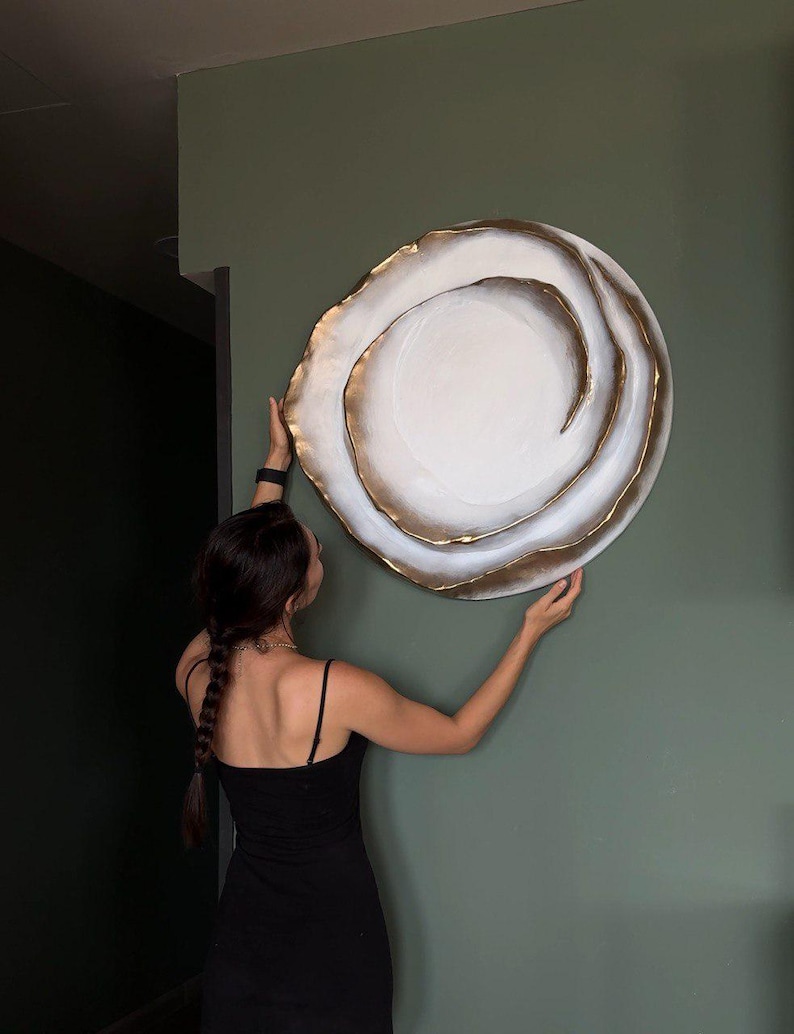 May include: A large, round wall art piece with a white center and gold-colored, layered edges. The artwork is being held against a muted green wall. The piece has a spiral design and a textured appearance.