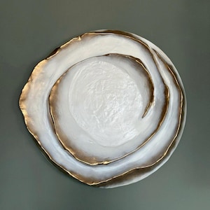 May include: A decorative, round, white plate with a gold-colored rim. The plate has a layered design with a textured surface. The gold rim adds a touch of elegance to the overall design. The plate is a decorative item.