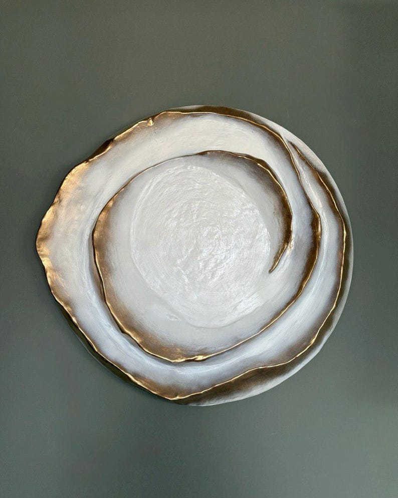 May include: A decorative, round, white plate with a gold-colored rim. The plate has a layered design with a textured surface. The gold rim adds a touch of elegance to the overall design. The plate is a decorative item.