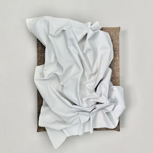 Può includere: Opera d'arte astratta con un tessuto bianco sgualcito drappeggiato su una tela rettangolare di juta marrone. Il tessuto è stratificato e strutturato, creando ombre e profondità. L'opera è montata su una parete bianca.