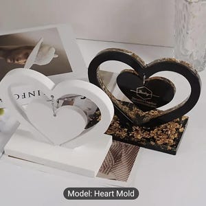 Pode incluir: Dois moldes decorativos em forma de coração. Um é branco, com um coração menor suspenso dentro. O outro é preto com flocos de ouro e um coração menor dentro. A base de cada molde é retangular. O texto "Model: Heart Mold" está na parte inferior.