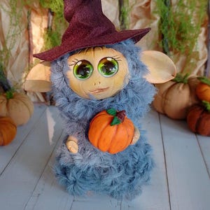 Peut inclure: Une poupée de créature d'Halloween faite à la main, avec un corps bleu duveteux, de grands yeux verts et un chapeau de sorcière bordeaux. Elle tient une petite citrouille orange. La poupée a de grandes oreilles et se trouve sur un fond de citrouilles et de feuillage d'automne.