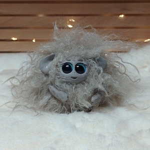 May include: A soft, grey toy creature with big, blue eyes and a smiling expression. The toy has fluffy, shaggy fur and small ears. It is on a white, fluffy surface, with a wooden backdrop and fairy lights.