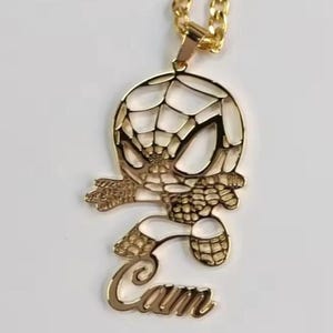 May include: Gold-toned pendant necklace featuring a stylized Spider-Man design. The pendant includes the name "Cam" in a cursive font. The necklace has a curb chain.