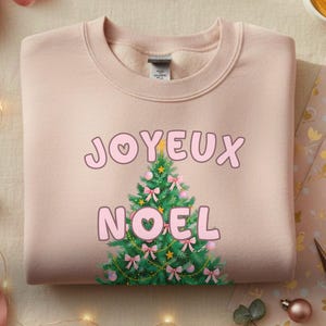以下が含まれることがあります： 「JOYEUX NOEL」の文字とクリスマスツリーのグラフィックがプリントされた、薄いピンク色のスウェットシャツ。 ピンクのリボン、オーナメント、ラッピングされたギフト、ラテアートが施されたコーヒーカップが写っています。