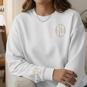 Puede incluir: Una sudadera blanca con iniciales bordadas en oro y un diseño a juego en la manga. La imagen muestra un hilo dorado y una opción de sudadera blanca.