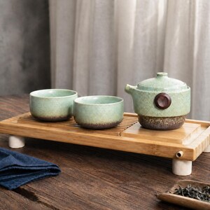 May include: A light green ceramic tea set with brown detailing. The set comprises a teapot with a wooden button, two small bowls, and a bamboo tray. The tray is supported by four white cylindrical legs. A blue cloth is on the wooden table.