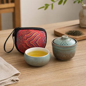 以下が含まれることがあります： お茶のセット。小さな丸い水色の陶器のカップに紅茶が入っており、お揃いの蓋付きのティーボウル、黒いジッパーとハンドルが付いた赤い模様のキャリングケースがあります。セットは木製のテーブルの上にあります。