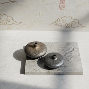 May include: Two small, metallic vases with a textured surface and a narrow opening. The vases are placed on a gray, rectangular stone. The background features a cream-colored wall with gold cloud designs.