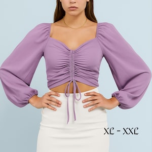 May include: A lilac crop top with long, voluminous sleeves and a square neckline. The top has a ruched front with a drawstring detail. The text "XL - XXL" is visible. The model is wearing a white skirt. The top is a fashionable garment.