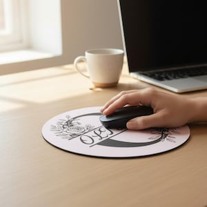 May include: A round, pink mousepad with a floral design and the name "Olivia" in black script. A black computer mouse rests on the pad, with a person's hand holding it. A laptop, mug, and plant are in the background.