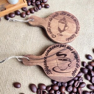 May include: Two wooden coffee-themed coasters with handles and string loops. One coaster features a coffee drink illustration and the text "Ce n'est pas du café, c'est de la motivation liquide." The other coaster has a coffee cup illustration and the text "L'élégance commence par une gorgée de café."