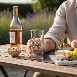 Peut inclure: Un verre transparent avec le texte "L'Apéro De PAPA" est présenté. Une bouteille de vin rosé, un petit bol de pistaches et une planche en bois avec du fromage, des raisins et des olives sont sur une table en bois.