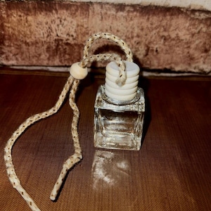 Może przedstawiać: Mała, kwadratowa szklana butelka z białą, warstwową drewnianą nakrętką i beżowym sznurkiem. Butelka to prawdopodobnie odświeżacz powietrza lub dyfuzor. Sznurek ma drewnianą koralik.