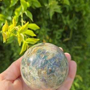 May include: A polished spherical stone featuring a complex mix of earthy brown, muted green, and vibrant magenta inclusions. The surface displays a natural, cracked texture with varying levels of opacity and light reflection throughout the orb.
