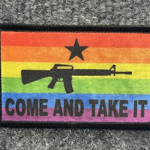 Puede incluir: Un parche rectangular con borde negro que presenta un fondo de bandera arcoíris horizontal. Una silueta negra de un rifle está centrada sobre el texto negro COME AND TAKE IT, con una estrella negra de cinco puntas sobre el rifle.