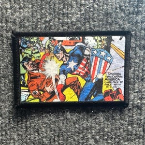 May include: A rectangular fabric patch featuring a comic book illustration of Captain America punching a figure in an orange uniform. The text reads: SMASHING THRU CAPTAIN AMERICA CAME FACE TO FACE WITH HITLER. The patch has a thick black border.