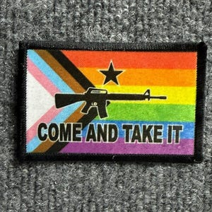 May include: A rectangular embroidered patch featuring a rainbow pride flag background with a black star and a black rifle silhouette in the center. The text COME AND TAKE IT is printed in bold black capital letters along the bottom edge.