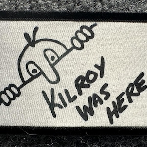 Op de afbeelding: Een rechthoekige stoffen patch met een witte achtergrond en een zwarte geborduurde rand. Het ontwerp toont een zwarte lijntekening van het Kilroy-figuur dat over een muur kijkt, vergezeld van de zwarte geborduurde tekst KILROY WAS HERE.