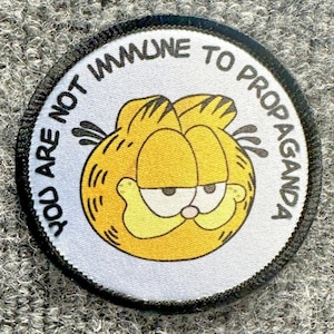 May include: A circular embroidered patch with a white background and a black stitched border. The center features a yellow cartoon cat face with black stripes and a skeptical expression. The text YOU ARE NOT IMMUNE TO PROPAGANDA is printed.