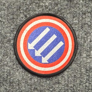 May include: A circular embroidered patch featuring a central blue field with three white diagonal arrows pointing downward to the left. The design is surrounded by concentric rings of white and red, finished with a black stitched border edge.