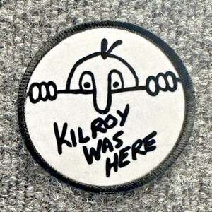 May include: A circular embroidered patch with a white background and a black border. The design features the classic Kilroy cartoon character peering over a horizontal line, with the black text KILROY WAS HERE printed below the character.