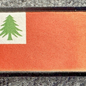 May include: A rectangular embroidered patch featuring a solid orange field with a white square in the upper left corner. Inside the white square is a green pine tree icon. The entire patch is bordered by a thick, black stitched edge.