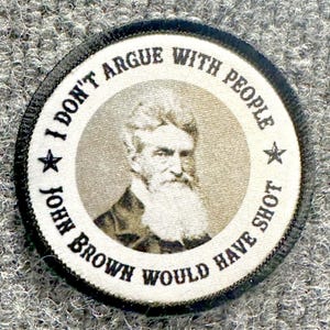 May include: A circular embroidered patch featuring a sepia-toned portrait of John Brown in the center. The white border contains the black text "I DON'T ARGUE WITH PEOPLE" at the top and "JOHN BROWN WOULD HAVE SHOT" at the bottom, separated by stars.