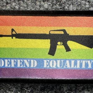 Pode incluir: Um emblema retangular com borda preta apresentando um fundo de bandeira arco-íris horizontal. Uma silhueta preta de um rifle tipo AR-15 está centralizada no meio. O texto branco "DEFEND EQUALITY" está impresso em negrito na parte inferior.