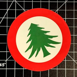 May include: A circular sticker featuring a solid green cedar tree silhouette centered on a white background, enclosed by a bold red circular border. The design is clean and graphic, presenting a stylized representation of a coniferous tree.