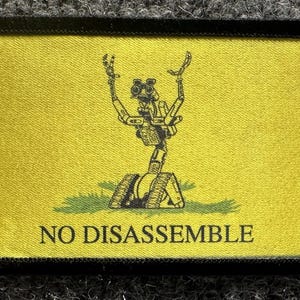 May include: A rectangular yellow patch with a black border featuring a black graphic of a robot with treads and raised arms standing on a patch of green grass. The text NO DISASSEMBLE is printed in black capital letters at the bottom of the patch.