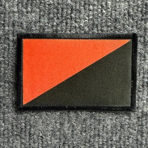 May include: A rectangular embroidered patch featuring a diagonal split design. The upper left triangle is bright red, and the lower right triangle is black. The entire patch is bordered by a thick, black stitched edge, creating a clean finish.
