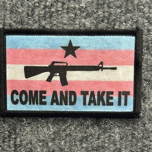 May include: A rectangular patch with a black border featuring a horizontal striped background in light blue, pink, and white. A black silhouette of a rifle is centered below a black star, with the black text COME AND TAKE IT printed below.