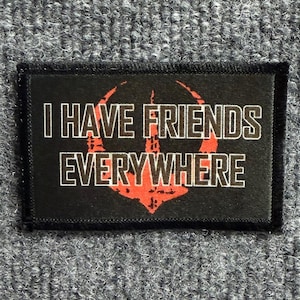 May include: A rectangular black patch with a stitched border featuring the white text I HAVE FRIENDS EVERYWHERE in a bold sans-serif font. Behind the text is a stylized red graphic symbol that resembles a circular emblem with a central point.