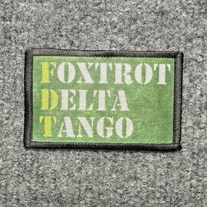May include: A rectangular morale patch with a green background and a black stitched border. The text FOXTROT DELTA TANGO is printed in white, with the letters F, D, and T highlighted in a pale yellow color, creating a vertical alignment.