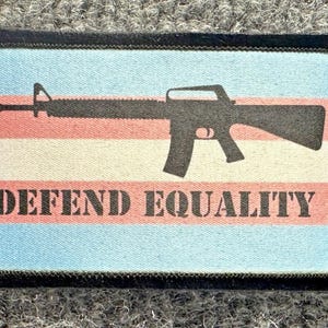 May include: A rectangular fabric patch with a black stitched border. The background features horizontal stripes of light blue, pink, and white. A black silhouette of a rifle is centered above the black text that reads DEFEND EQUALITY.