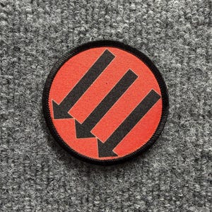 Puede incluir: Un parche bordado circular que presenta un fondo rojo vibrante con tres flechas negras audaces que apuntan diagonalmente hacia abajo a la izquierda. El parche está rematado con un borde grueso cosido en negro alrededor del borde exterior.