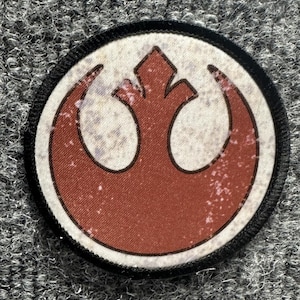May include: A circular embroidered patch featuring a rust-colored Rebel Alliance starbird symbol centered on a distressed off-white background. The design is enclosed by a thick black stitched border, creating a clean and defined edge for the emblem.