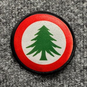 May include: A circular embroidered patch featuring a green cedar tree centered on a white background, surrounded by a thick red border and finished with a black stitched edge. The design is clean and symmetrical, showcasing a classic tree emblem.